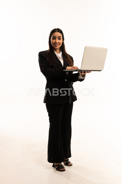 استخدام اللابتوب في تنظيم خطط العمل، استخدام الأجهزة التقنية الحديثة في إنجاز المهام، بورتريه لامرأة عربية سعودية خليجية مبتسمة ترتدي البدلة الرسمية السوداء تقف باستقامة و تستخدم الحاسب المحمول, خلفية بيضاء
