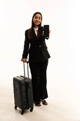 تطبيقات الهاتف الذكي، الاستمتاع بالرحلات السياحية في وقت العطلة, بورتريه لامرأة عربية سعودية خليجية مبتسمة ترتدي البدلة الرسمية السوداء تقف باستقامة و تقوم بجر حقيبة الامتعة و تقوم بعرض شاشة الموبايل, خلفية بيضاء
