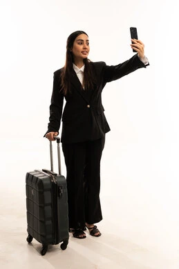 التقاط صورة سيلفي عبر الهاتف المحمول، الذهاب في رحلة سفر، الفرح لقضاء رحلة صيفية ممتعة، بورتريه لامرأة عربية سعودية خليجية ترتدي البدلة الرسمية السوداء تقف باستقامة و تقوم بجر حقيبة الامتعة و تحمل بيدها الموبايل, ايماءات السعادة والسرور، خلفية بيضاء