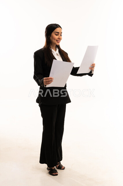 Reviewing notes, preparing work reports, a portrait of a Saudi Arabian woman from the Gulf region wearing a black formal suit, standing upright and holding a stack of papers, reviewing and following up on plans and projects, white background