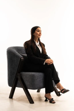 Sitting confidently and upright, paying attention to elegance and appearance, a portrait of a Saudi Arabian Gulf woman wearing a black formal suit, sitting in a comfortable chair and looking away with pride, against a white background.