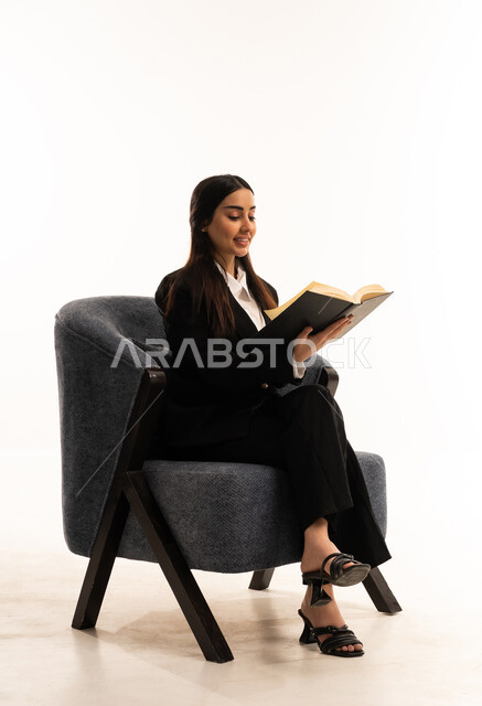 Spending time reading, learning, and self-educating: A portrait of a Saudi Arabian woman from the Gulf region wearing a black formal suit, sitting in a comfortable chair, holding a book, enjoying reading books, stories, and novels; a culture of self-learning; white background.