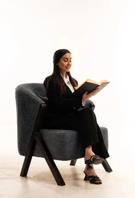 Spending time reading, learning, and self-educating: A portrait of a Saudi Arabian woman from the Gulf region wearing a black formal suit, sitting in a comfortable chair, holding a book, enjoying reading books, stories, and novels; a culture of self-learning; white background.