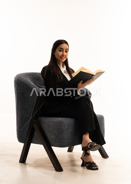 الاستمتاع بمطالعة الكتب والقصص والروايات, قضاء الوقت بالقراءة والتعلم والتثقف، بورتيه لامرأة عربية سعودية خليجية ترتدي البدلة الرسمية السوداء تجلس على كرسي مريح و تحمل بيدها  كتاب، ثقافة التعلم الذاتي, خلفية بيضاء