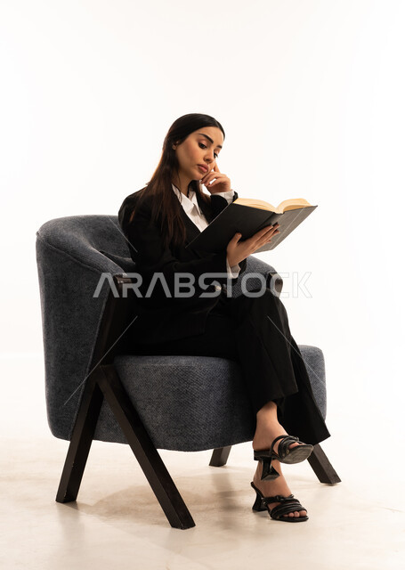 هواية القراءة والاستكشاف، الاستمتاع بالكتب والقصص والروايات، بورتيه لامرأة عربية سعودية خليجية ترتدي البدلة الرسمية السوداء تجلس على كرسي مريح و تحمل بيدها  كتاب، ثقافة التعلم الذاتي, خلفية بيضاء