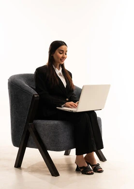 The concept of online employment, browsing social media on a laptop, remote work via the internet, integrating technology into working life; a portrait of a Saudi Arabian woman from the Gulf region wearing a black formal suit, sitting in a comfortable chair and using a laptop, against a white background.