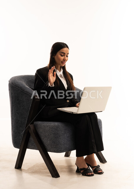 A remote business meeting, online chat and communication, using modern and advanced devices and technologies; a portrait of a Saudi Arabian Gulf woman wearing a black formal suit, sitting in a comfortable chair and using a laptop, white background.