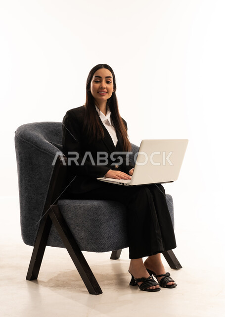 دمج التكنولوجيا في الحياة العملية, تصفح مواقع التواصل الاجتماعي بواسطة الحاسب المحمول، العمل عن بعد عبر الانترنت، بورتيه لامرأة عربية سعودية خليجية ترتدي البدلة الرسمية السوداء تجلس على كرسي مريح و تقوم باستخدام جهاز اللابتوب، خلفية بيضاء