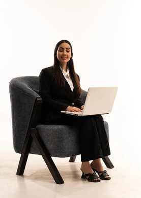 دمج التكنولوجيا في الحياة العملية, تصفح مواقع التواصل الاجتماعي بواسطة الحاسب المحمول، العمل عن بعد عبر الانترنت، بورتيه لامرأة عربية سعودية خليجية ترتدي البدلة الرسمية السوداء تجلس على كرسي مريح و تقوم باستخدام جهاز اللابتوب، خلفية بيضاء