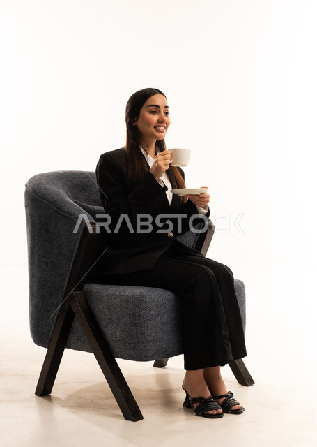 قضاء وقت الفراغ باحتساء المشروب المفضل, الجلوس والاستمتاع بالمشروبات الساخنة والأعشاب الشتوية الدافئة, بورتيه لامرأة عربية سعودية خليجية ترتدي البدلة الرسمية السوداء تجلس على كرسي مريح و تحمل بيدها فنجان من الشاي، خلفية بيضاء