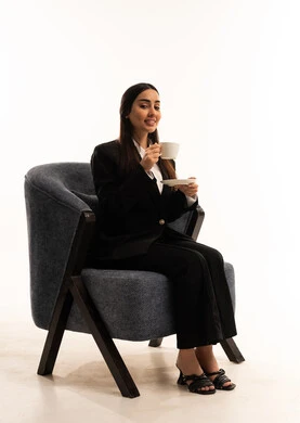 Sit back and enjoy hot drinks and warm winter herbs, spend your free time sipping your favorite beverage. Portrait of a Saudi Arabian Gulf woman wearing a black formal suit, sitting in a comfortable chair and holding a cup of tea, white background