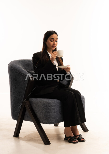 Spending a pleasant and quiet time, sitting and enjoying hot drinks and warm winter herbs, sipping your favorite beverage. Portrait of a Saudi Arabian Gulf woman wearing a black formal suit, sitting in a comfortable chair and holding a cup of tea, white background.