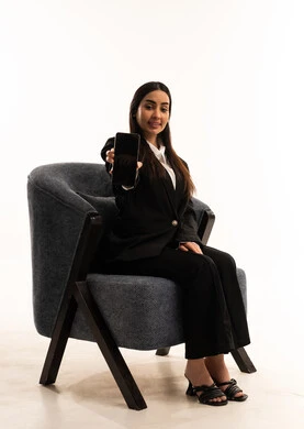 Displaying a blank black screen on a mobile phone, using advanced modern technology, accomplishing work tasks remotely, a modern and advanced technological device, a portrait of a Saudi Arabian Gulf woman wearing a black formal suit, sitting in a comfortable chair and holding a mobile phone, white background