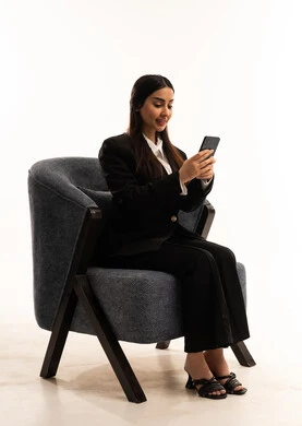 العمل عن بعد عبر الجوال، ايماءات الاندماج والتركيز، استخدام التقنية الحديثه المتطورة في الحياة العملية، بورتيه لامرأة عربية سعودية خليجية ترتدي البدلة الرسمية السوداء تجلس على كرسي مريح و تحمل بيدها موبايل, خلفية بيضاء