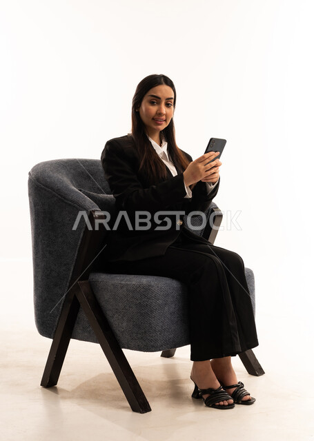 تصفح مواقع التواصل الاجتماعي, العمل عن بعد عبر الجوال، النظر الى الكاميرا بايماءات السعادة، استخدام التقنية الحديثه المتطورة في الحياة العملية، بورتيه لامرأة عربية سعودية خليجية ترتدي البدلة الرسمية السوداء تجلس على كرسي مريح و تحمل بيدها موبايل, خلفية بيضاء
