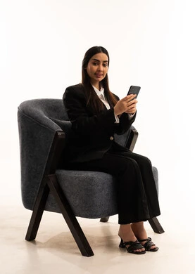 تصفح مواقع التواصل الاجتماعي, العمل عن بعد عبر الجوال، النظر الى الكاميرا بايماءات السعادة، استخدام التقنية الحديثه المتطورة في الحياة العملية، بورتيه لامرأة عربية سعودية خليجية ترتدي البدلة الرسمية السوداء تجلس على كرسي مريح و تحمل بيدها موبايل, خلفية بيضاء