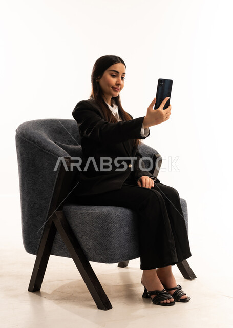 التواصل والدردشة مع الأهل والأصدقاء عبر تطبيقات الجوال,  إجراء مكالمات الفيديو عبر الانترنت، بورتيه لامرأة عربية سعودية خليجية ترتدي البدلة الرسمية السوداء تجلس على كرسي مريح و تحمل بيدها موبايل, خلفية بيضاء