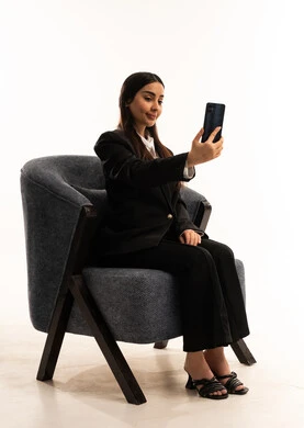التواصل والدردشة مع الأهل والأصدقاء عبر تطبيقات الجوال,  إجراء مكالمات الفيديو عبر الانترنت، بورتيه لامرأة عربية سعودية خليجية ترتدي البدلة الرسمية السوداء تجلس على كرسي مريح و تحمل بيدها موبايل, خلفية بيضاء