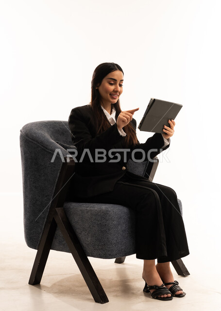 إستخدام اجهزة تقنية حديثة، إيماءات السعادة والسرور، بورتيه لامرأة عربية سعودية خليجية ترتدي البدلة الرسمية السوداء تجلس على كرسي مريح و تحمل بيدها الجهاز اللوحي، الاعتناء بالمظهر الخارجي، خلفية بيضاء