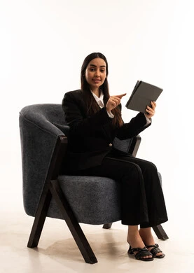 Using tablet apps and programs, gestures of happiness and joy, a portrait of a Saudi Arabian Gulf woman wearing a black formal suit, sitting in a comfortable chair and holding a tablet, taking care of her appearance, white background