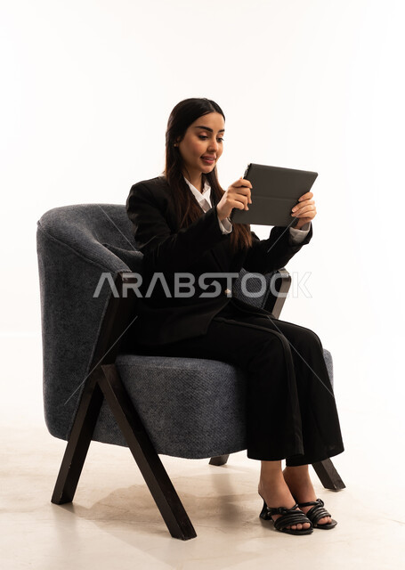 مفهوم إدارة المشاريع عن بعد, إستخدام اجهزة تقنية حديثة، إيماءات السعادة والسرور، بورتيه لامرأة عربية سعودية خليجية ترتدي البدلة الرسمية السوداء تجلس على كرسي مريح و تحمل بيدها الجهاز اللوحي، الاعتناء بالمظهر الخارجي، خلفية بيضاء