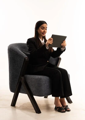 مفهوم إدارة المشاريع عن بعد, إستخدام اجهزة تقنية حديثة، إيماءات السعادة والسرور، بورتيه لامرأة عربية سعودية خليجية ترتدي البدلة الرسمية السوداء تجلس على كرسي مريح و تحمل بيدها الجهاز اللوحي، الاعتناء بالمظهر الخارجي، خلفية بيضاء