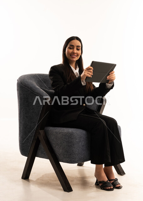 Completing work tasks via tablet, gestures of happiness and joy, a portrait of a Saudi Arabian Gulf woman wearing a black formal suit, sitting in a comfortable chair and holding a tablet, taking care of her appearance, white background