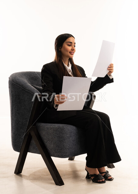 Using white paper to record important notes and information; tools for writing down daily tasks; gestures of happiness and optimism; a portrait of a Saudi Arabian woman wearing a black formal suit, sitting in a comfortable chair, holding a stack of papers; reviewing and following up on plans and projects; white background.