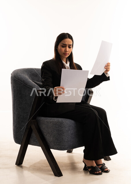 أدوات تدوين المهام اليومية، استخدام الورق الأبيض في تسجيل الملاحظات والمعلومات المهمة,  إيماءات التركيز والتمعن، بورتيه لامرأة عربية سعودية خليجية ترتدي البدلة الرسمية السوداء تجلس على كرسي مريح و تحمل بيدها مجموعة من الاوراق، مراجعة و متابعة الخطط و المشاريع, خلفية بيضاء