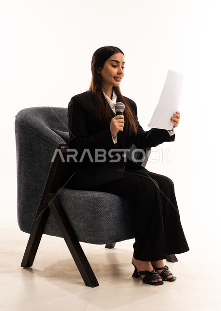 Effective communication and dialogue, conducting interviews and asking questions, the concept of art and its positive impact, a portrait of a smiling Saudi Arabian woman wearing a black formal suit, sitting in a comfortable chair, holding a microphone and a piece of paper, attention to appearance, white background