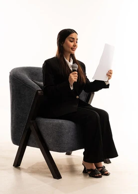 Effective communication and dialogue, conducting interviews and asking questions, the concept of art and its positive impact, a portrait of a smiling Saudi Arabian woman wearing a black formal suit, sitting in a comfortable chair, holding a microphone and a piece of paper, attention to appearance, white background