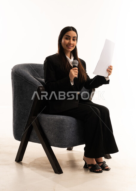 Conducting interviews and asking questions, gestures and expressions of integration and interaction, the concept of art and its positive impact, a portrait of a smiling Saudi Arabian woman wearing a black formal suit, sitting in a comfortable chair, holding a microphone and a piece of paper, attention to appearance, white background