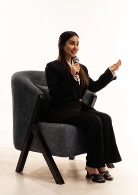 Gestures and expressions of integration and interaction; the concept of art and its positive impact; a portrait of a Saudi Arabian Gulf woman wearing a black formal suit, sitting in a comfortable chair and holding a microphone; attention to outward appearance; white background.