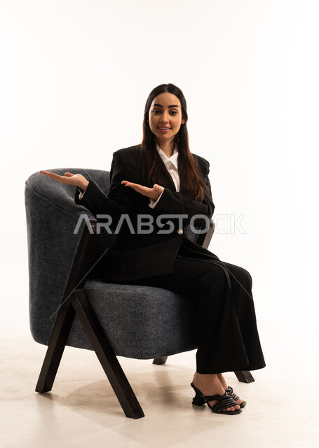 إشارات يد تدل على الترحيب, مفهوم الأناقة والاعتناء بالمظهر الخارجي، بورتريه لامرأة عربية سعودية خليجية ترتدي البدلة الرسمية السوداء تجلس على كرسي مريح, ابتسامة لطيفة تدل على المحبة، خلفية بيضاء