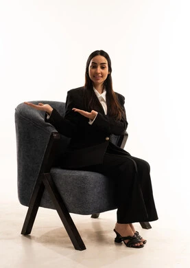 Hand gestures conveying welcome, a sense of elegance and attention to appearance, a portrait of a Saudi Arabian woman from the Gulf region wearing a black formal suit, seated in a comfortable chair, a kind smile conveying affection, white background