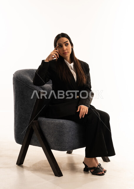 Deep thinking and absentmindedness, trying to find a solution to difficult problems, anxiety and confusion in making the right decision, a portrait of a Saudi Arabian Gulf woman wearing a black formal suit, sitting in a comfortable chair with her hand on her head in thoughtful gestures, concerned with her appearance, white background