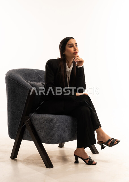 القلق والحيرة في اتخاذ القرار الصحيح، التفكير بعمق وشرود الذهن، محاولة إيجاد حل للمشكلات الصعبة، بورتريه لامرأة عربية سعودية خليجية ترتدي البدلة الرسمية السوداء تجلس على كرسي مريح و تضع يدها على ذقنها بايماءات التفكير, مفهوم الأناقة والاعتناء بالمظهر الخارجي، خلفية بيضاء
