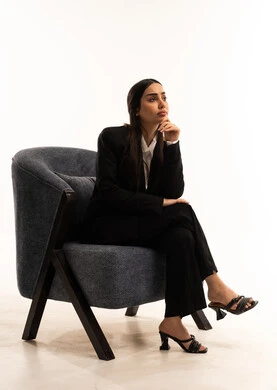 Anxiety and hesitation in making the right decision, deep thinking and absentmindedness, trying to find a solution to difficult problems; a portrait of a Saudi Arabian Gulf woman wearing a black formal suit, sitting in a comfortable chair with her hand on her chin in thoughtful gestures; the concept of elegance and attention to outward appearance; white background.