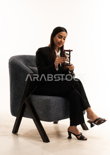 القيام بمراقبة توقيت الرمل, تحديد أوقات و مواعيد العمل، مفهوم الأناقة والاعتناء بالمظهر الخارجي، بورتريه لامرأة عربية سعودية خليجية ترتدي البدلة الرسمية السوداء تجلس على كرسي مريح و تمسك بيدها ساعة رملية, الفكر الذهني العميق, خلفية بيضاء