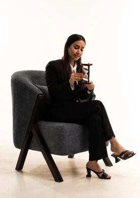 Monitoring the sand, setting work times and appointments, the concept of elegance and attention to outward appearance, a portrait of a Saudi Arabian Gulf woman wearing a black formal suit, sitting in a comfortable chair and holding an hourglass, deep mental contemplation, white background