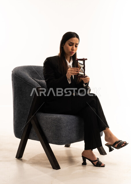 تحديد أوقات و مواعيد العمل، مفهوم الأناقة والاعتناء بالمظهر الخارجي، بورتريه لامرأة عربية سعودية خليجية ترتدي البدلة الرسمية السوداء تجلس على كرسي مريح و تمسك بيدها ساعة رملية, الفكر الذهني العميق, خلفية بيضاء