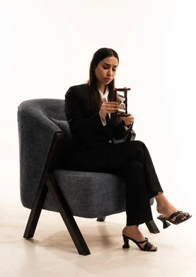 تحديد أوقات و مواعيد العمل، مفهوم الأناقة والاعتناء بالمظهر الخارجي، بورتريه لامرأة عربية سعودية خليجية ترتدي البدلة الرسمية السوداء تجلس على كرسي مريح و تمسك بيدها ساعة رملية, الفكر الذهني العميق, خلفية بيضاء