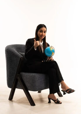 عرض مجسم الكرة الأرضية, مفهوم الأناقة والاعتناء بالمظهر الخارجي، ايماءات السعادة و السرور, بورتريه لامرأة عربية سعودية خليجية ترتدي البدلة الرسمية السوداء تجلس على كرسي مريح و ترفع اصبع الابهام باشارة اللايك, البحث والإستكشاف حول العالم، خلفية بيضاء