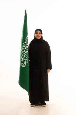 يوم العلم 11 مارس، مفهوم حب الوطن والانتماء اليه، ذكرى اليوم الوطني السعودي 23 سبتمبر، بورتريه لسيدة عربية خليجية سعودية محجبة مبتسمة ترتدي عباءة سوداء تحمل بيدها علم المملكة وتنظر الى الكاميرا بإيماءات السرور والثقة بالنفس، صورة الجسم بالكامل، خلفية بيضاء