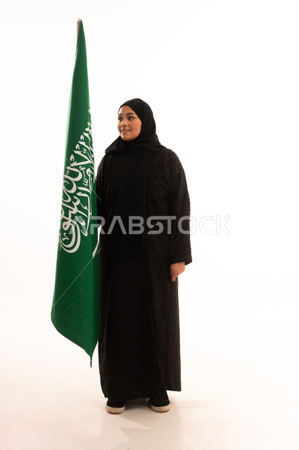 النظر إلى العلم بايماءات الاعتزاز والفخربالانتماء إلى الوطن، ذكرى اليوم الوطني السعودي 23 سبتمبر، يوم العلم 11 مارس، إظهار الحب والولاء للبلاد، بورتريه لسيدة عربية خليجية سعودية محجبة ترتدي عباءة سوداء تحمل بيدها علم المملكة، صورة الجسم بالكامل، خلفية بيضاء