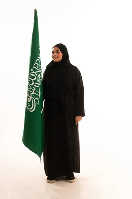 Looking at the flag with gestures of pride and belonging to the homeland, Saudi National Day, September 23, Flag Day, March 11, showing love and loyalty to the country, portrait of a veiled Saudi Arabian Gulf woman wearing a black abaya holding the flag of the Kingdom, full body image, white background