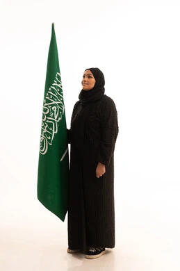 الوقوف باستقامة بايماءات الاعتزاز والفخربالانتماء إلى الوطن، ذكرى اليوم الوطني السعودي 23 سبتمبر، يوم العلم 11 مارس، إظهار الحب والولاء للبلاد، بورتريه لسيدة عربية خليجية سعودية محجبة ترتدي عباءة سوداء تحمل بيدها علم المملكة، صورة الجسم بالكامل، خلفية بيضاء