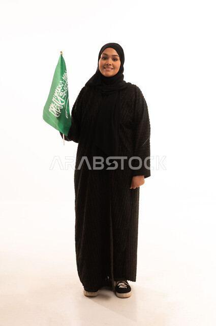 ذكرى اليوم الوطني السعودي 23 سبتمبر، يوم العلم 11 مارس، مفهوم حب الوطن والانتماء اليه، بورتريه لسيدة عربية خليجية سعودية محجبة مبتسمة ترتدي عباءة سوداء تحمل بيدها علم المملكة وتنظر الى الكاميرا بإيماءات السرور والثقة بالنفس، صورة الجسم بالكامل، خلفية بيضاء