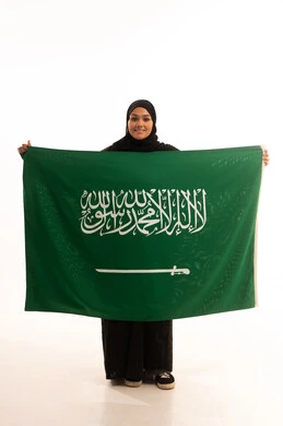 Flag Day, March 11, the concept of patriotism and belonging to the homeland, the memory of Saudi National Day, September 23, a portrait of a smiling veiled Saudi Arabian Gulf woman wearing a black abaya, holding the flag of the Kingdom in her hand and looking at the camera with gestures of pleasure and self-confidence, a full body image, a white background