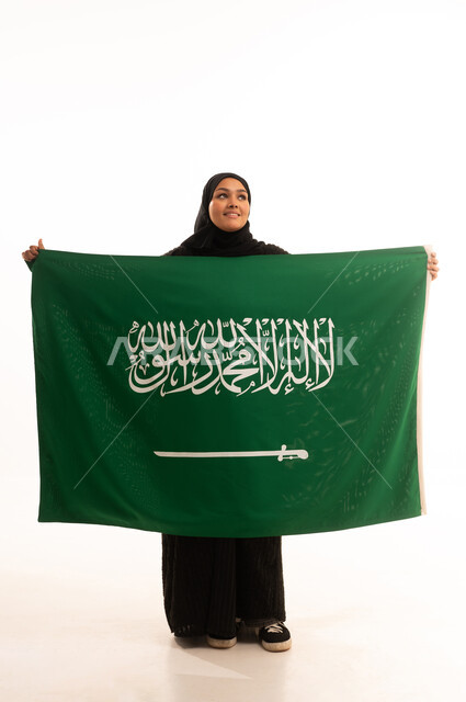 التطلع والنظر إلى الاعلى بايماءات الاعتزاز والفخربالانتماء إلى الوطن، ذكرى اليوم الوطني السعودي 23 سبتمبر، يوم العلم 11 مارس، إظهار الحب والولاء للبلاد، بورتريه لسيدة عربية خليجية سعودية محجبة ترتدي عباءة تحمل بيدها علم المملكة، صورة الجسم بالكامل، خلفية بيضاء